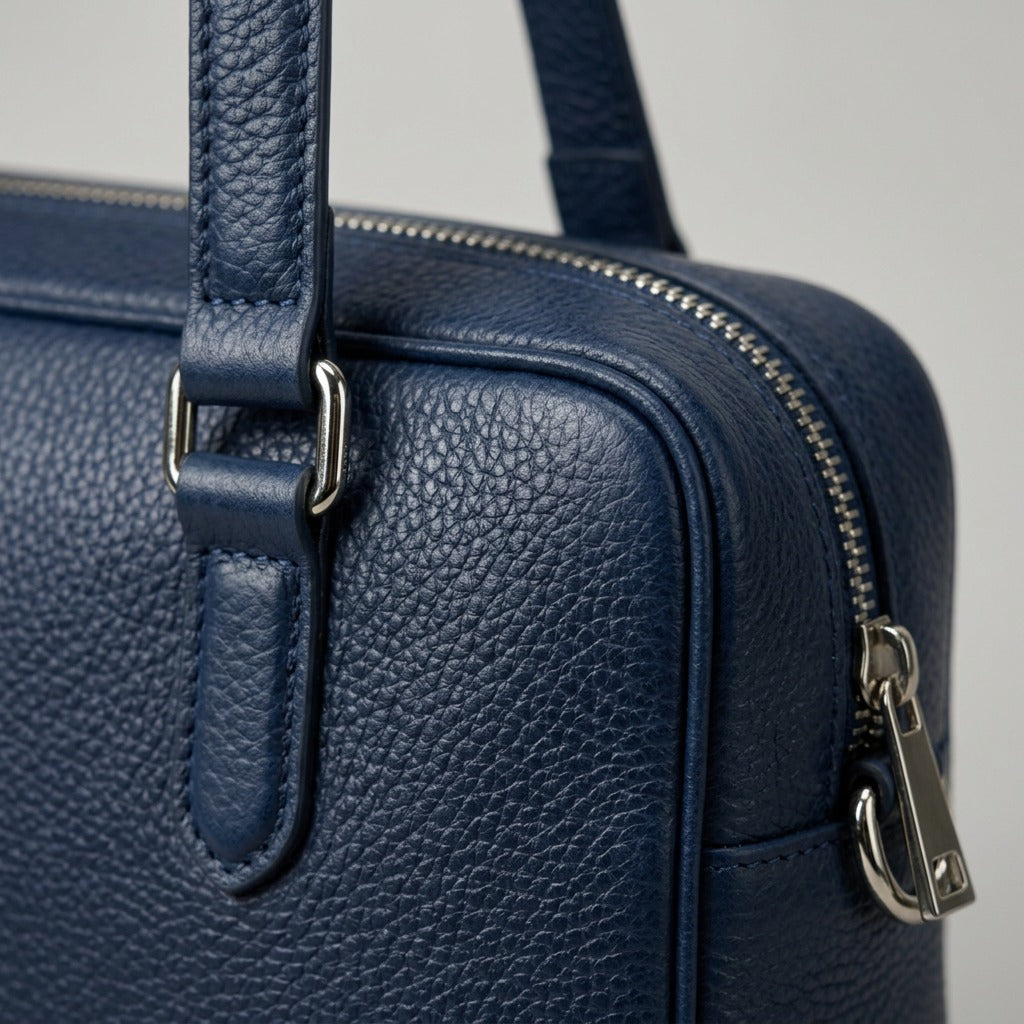A macro photograph showing the precise blue leather texture and silver buckle detail on the laptop bag.