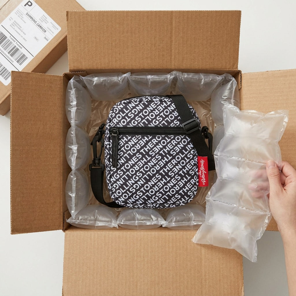 A top-down view of a cross-body bag nestled securely in a cardboard box, surrounded by protective air pillows for safe transit. The bag features a black background patterned repeatedly with "The Rolling Stones" in white text and has a prominent red fabric tag with "The Rolling Stones" logo attached. It fits precisely into the shipping enclosure.